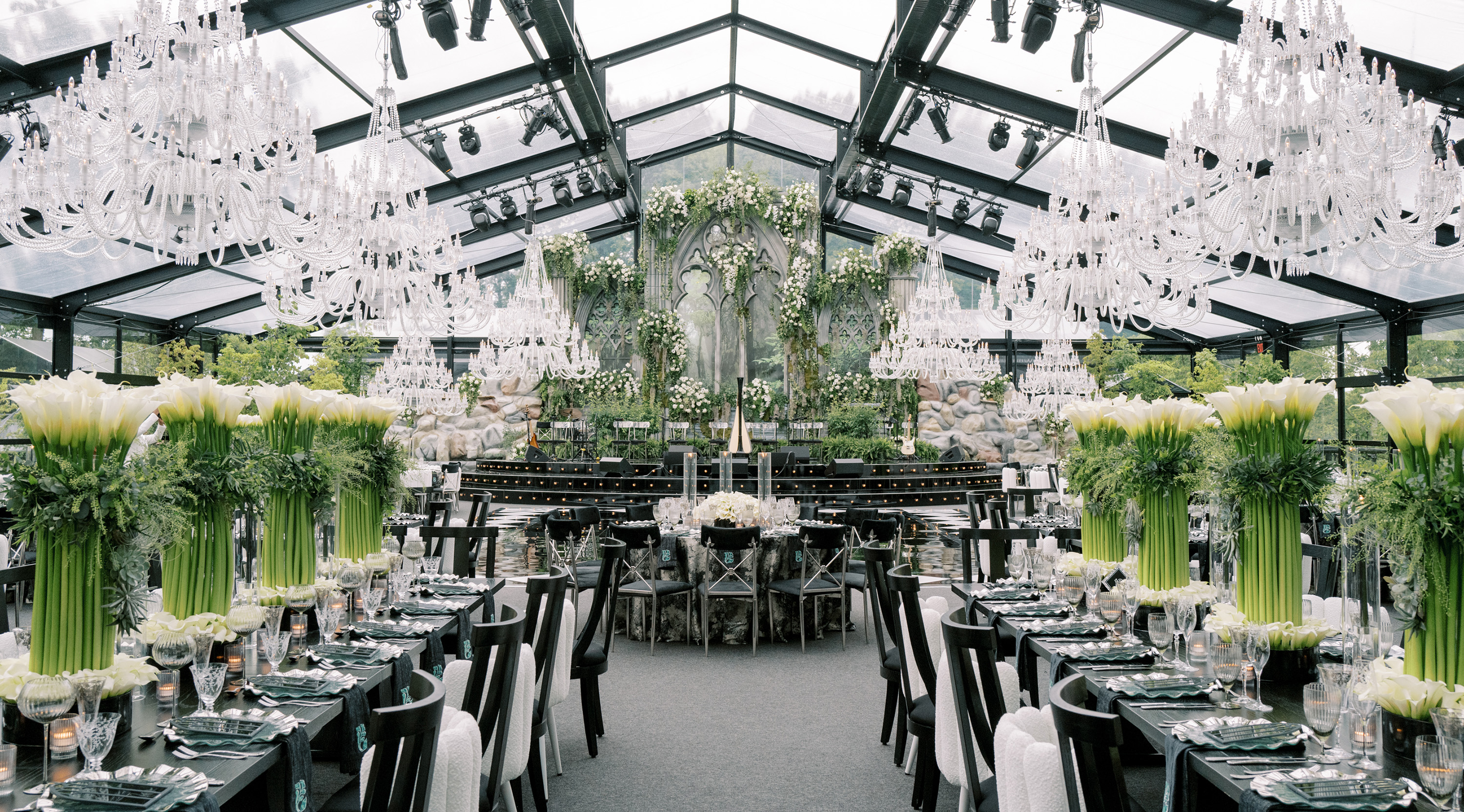 Green and white florals, crystal chandeliers, and black seating and accents enhance the decor of a glass tent set for a wedding dinner gala