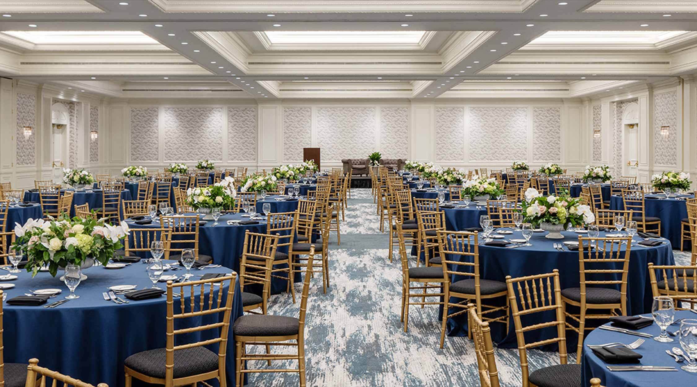 A ballroom venue with neutral walls and flooring decorated with blue tablecloths, florals, and gold chiavari chairs in the Marquis Ballroom at Nemacolin resort